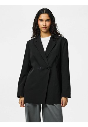 Vrouw met krullend donker haar, gekleed in een oversized zwarte blazer, wit topje en grijze broek met krijtstreep, staand tegen een witte achtergrond.