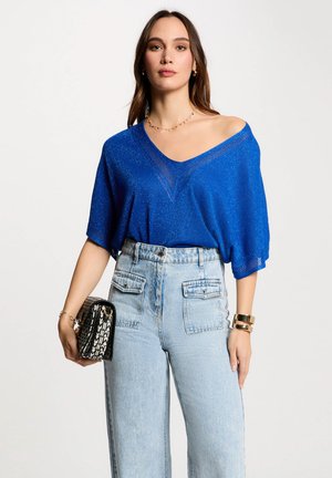 Blauwe, losvallende V-hals top gemaakt van gestructureerd, sprankelend materiaal, gecombineerd met hoog-wijde lichtblauwe denim jeans. Accessoires zijn onder andere een zwarte, gepatterneerde handtas.
