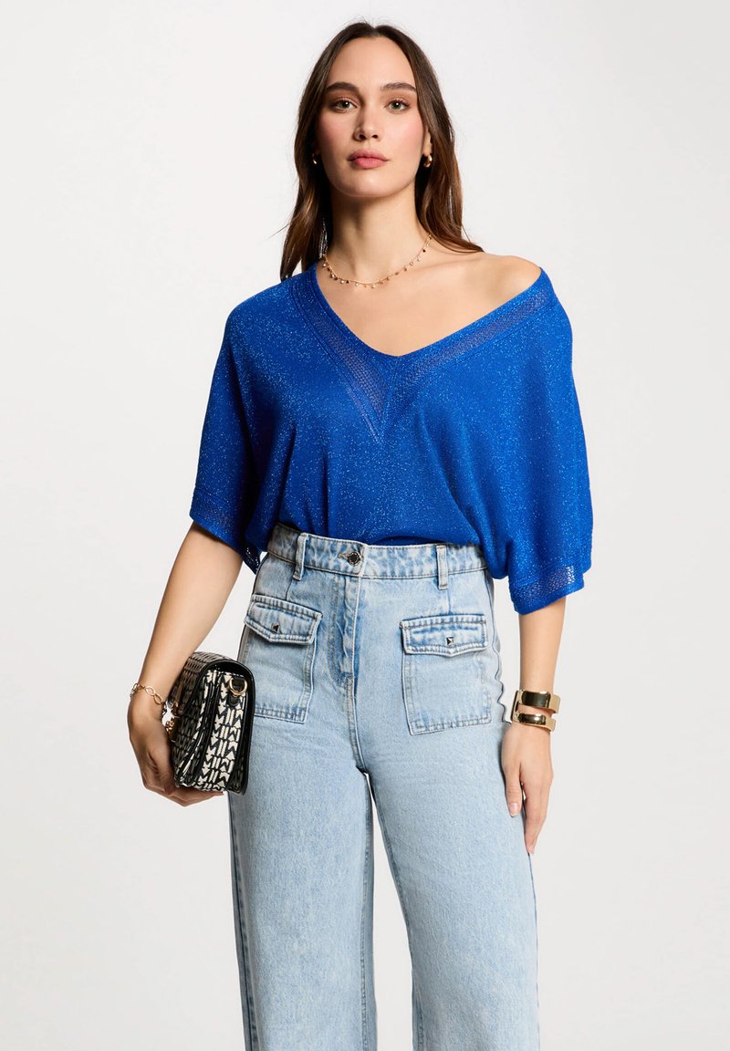 Lockere, blaue V-Ausschnitt-Top aus strukturiertem, glitzerndem Material, kombiniert mit hoch taillierten hellen Jeans. Als Accessoires wird eine schwarze, gemusterte Handtasche getragen.