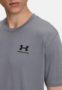 Siva kratka majica s kratkimi rokavi iz bombaža, ki ima ribast kroj okoli vratu in črn izvezeni logotip na levem prsnem delu.