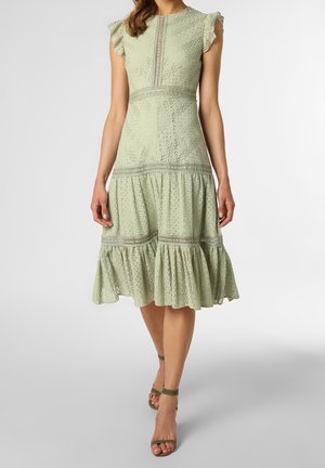 Femme portant une robe midi en dentelle œillets vert clair avec manches à volants et jupe à étages, associée à des talons à bride cheville vert olive.
