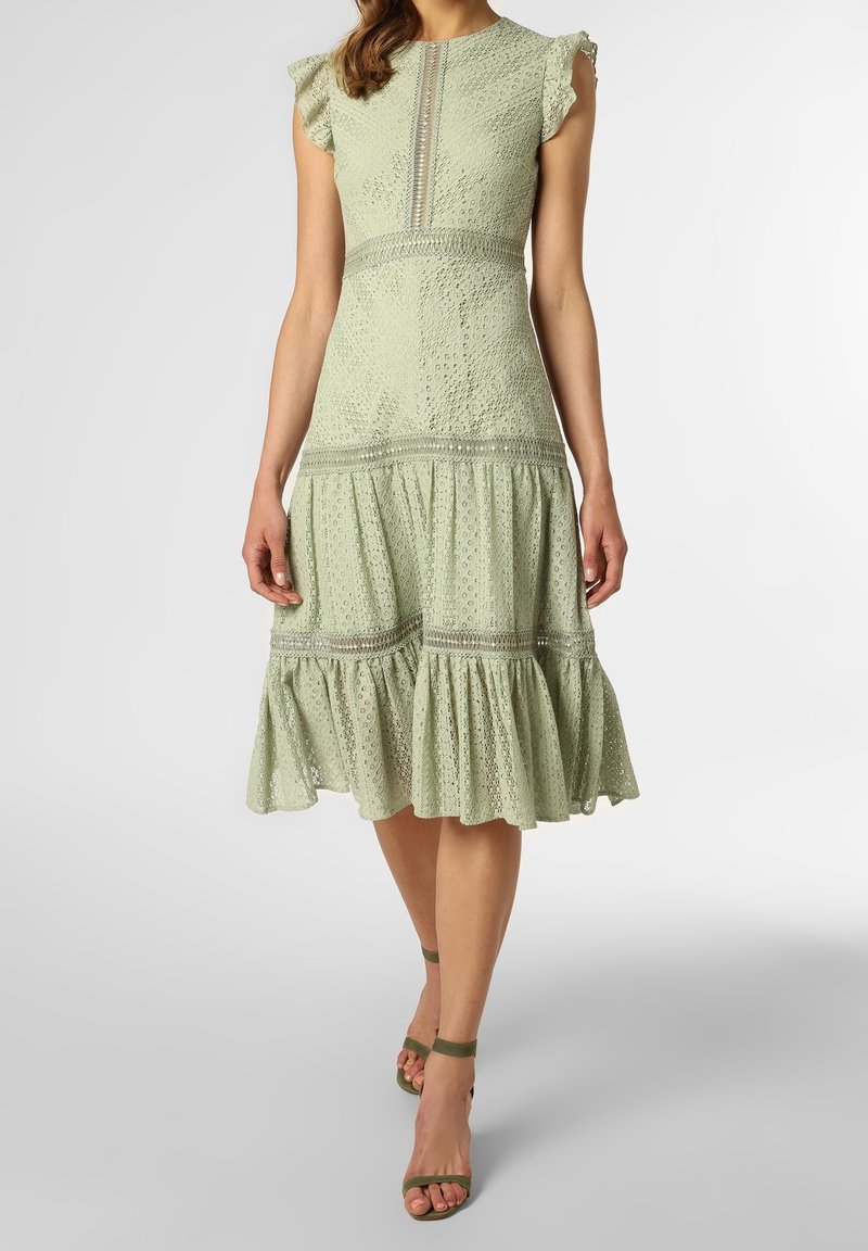 Femme portant une robe midi en dentelle œillets vert clair avec manches à volants et jupe à étages, associée à des talons à bride cheville vert olive.