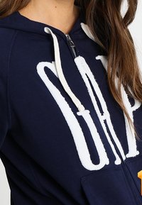 Marineblauwe zip-up hoodie met witte geborduurde "GAP" logo. Voorzien van een capuchon met trekkoord en zachte, gestructureerde stof.
