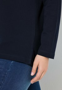Marineblaue Langarmbluse mit Rundhalsausschnitt, seitlichen Schlitzen und strukturiertem Stoff. Eine Hand ruht auf dem unteren Saum über Jeans.