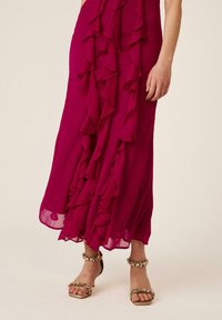 Mujer con vestido midi rosa oscuro con volantes combinado con tacones abiertos dorados con joyas, de pie sobre un fondo sencillo.