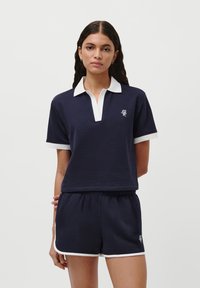 Námořnický modrý polo top s bílým límcem a manžetami, s vyšívaným logem. Měkká tkanina; volný střih. V kombinaci s odpovídajícími šortkami.
