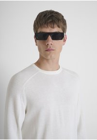 Zwarte rechthoekige zonnebril gedragen door een persoon, gecombineerd met een lichtbeige, gestructureerde ronde halstrui. De achtergrond is eenvoudig wit.