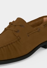 Mocassin en daim marron avec surpiqûres mocassin, détails à lacets et semelle en caoutchouc noir sur fond clair uni.