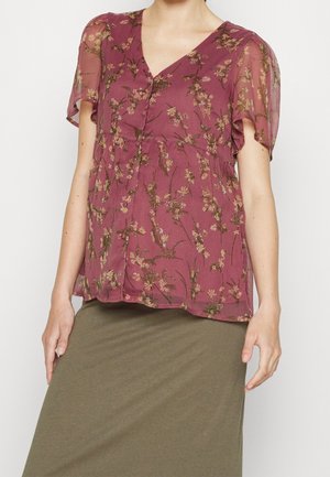 Kobieta ubrana w przezroczystą kwiatową bluzkę w kolorze mauve z krótkimi, falującymi rękawami i zapięciem na guziki z przodu, połączoną z oliwkową spódnicą.