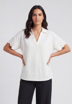 Femme aux cheveux foncés portant un chemisier blanc à manches courtes avec des manches festonnées à œillets et un pantalon noir, debout les mains sur les hanches.