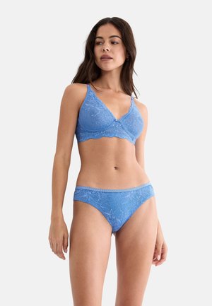 Kobieta w niebieskim koronkowym biustonoszu i majtkach, stojąca na tle jednolitego białego tła, patrząca lekko w lewo.