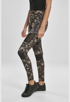 Personne portant des baskets noires et des leggings à motif camouflage avec des panneaux en mesh noir, debout devant un fond gris uni.
