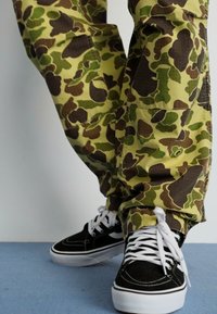 Vihreät, ruskeat ja keltaiset camo-housut, joissa on väljä istuvuus. Mustat lenkkarit, joissa on valkoisia yksityiskohtia ja nauhat. Littee kangaspinta.