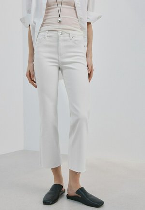 Person iført hvide korte jeans, beige ribbet top, hvid åben skjorte, sorte slip-on sko og en fløjtenøglering mod en ensfarvet baggrund.