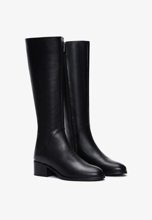 Une paire de bottes en cuir noir jusqu'au genou avec des talons bas et des fermetures éclair sur le côté, présentée sur un fond blanc.