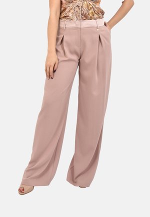 Donna che indossa pantaloni larghi rosa cipria a vita alta con pieghe, abbinati a una canotta fantasia e scarpe beige con punta aperta.