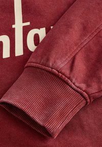 Donkerrode sweatshirt met geribbelde manchetten en een gladde textuur. Heeft een opvallend ontwerp met crèmekleurige tekst, met lichte vervaging voor een gedragen uitstraling.