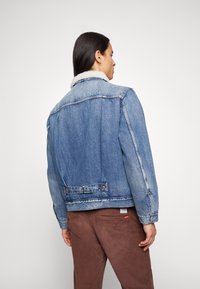 Denimjack met een lichtblauwe wassing, schapenkraag en twee borstzakken. Versleten details aan de zoom en manchetten. Bruine corduroy broek.