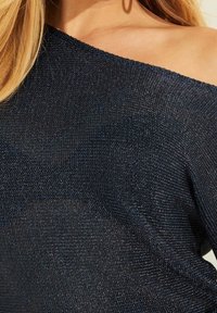 Dzianinowy ciemnoniebieski top o asymetrycznym, odkrywającym ramię kroju. Materiał ma subtelny połysk i teksturowany wzór z drobnymi metalicznymi włóknami.
