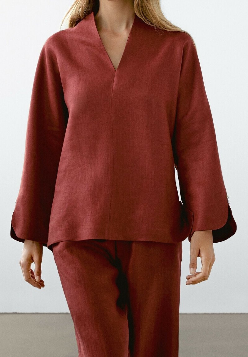 Femme portant un haut ample à manches longues rouge rouille et un pantalon assorti sur un fond clair uni.