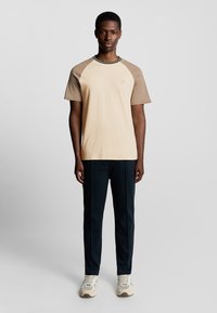 Beige og tan farveblokeret t-shirt med rund hals og korte ærmer, parret med mørkeblå bukser og lyse sneakers.