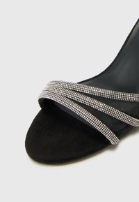 Juodos zomšinės aukštakulnės sandalai su kryžminėmis dirželių juostomis, puoštomis sidabro spalvos rhinestone akmenimis. Švelnios matinės apdailos su smailiu pirštu dizainu.