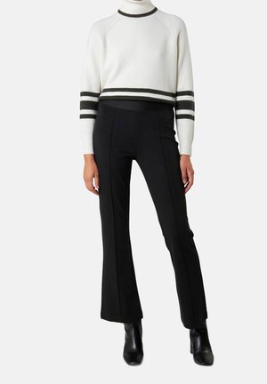 Nainen pukeutunut valkoiseen lyhennettyyn pooloneuleeseen, jossa on mustat raidat, mustiin istuviin liehuhousuihin ja mustiin nilkkureihin.