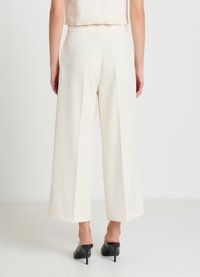 Pantalon ample de couleur crème avec plis à l'avant, texture lisse, taille haute et ourlet à la cheville, porté avec des talons noirs.