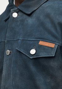 Κοντινό πλάνο τσέπης σκούρας μπλε σατέν μπουφάν με ασημί κουμπί τύπου snap της Calvin Klein και μικρή ταμπέλα από ταμπά δέρμα με το λογότυπο της εταιρείας.