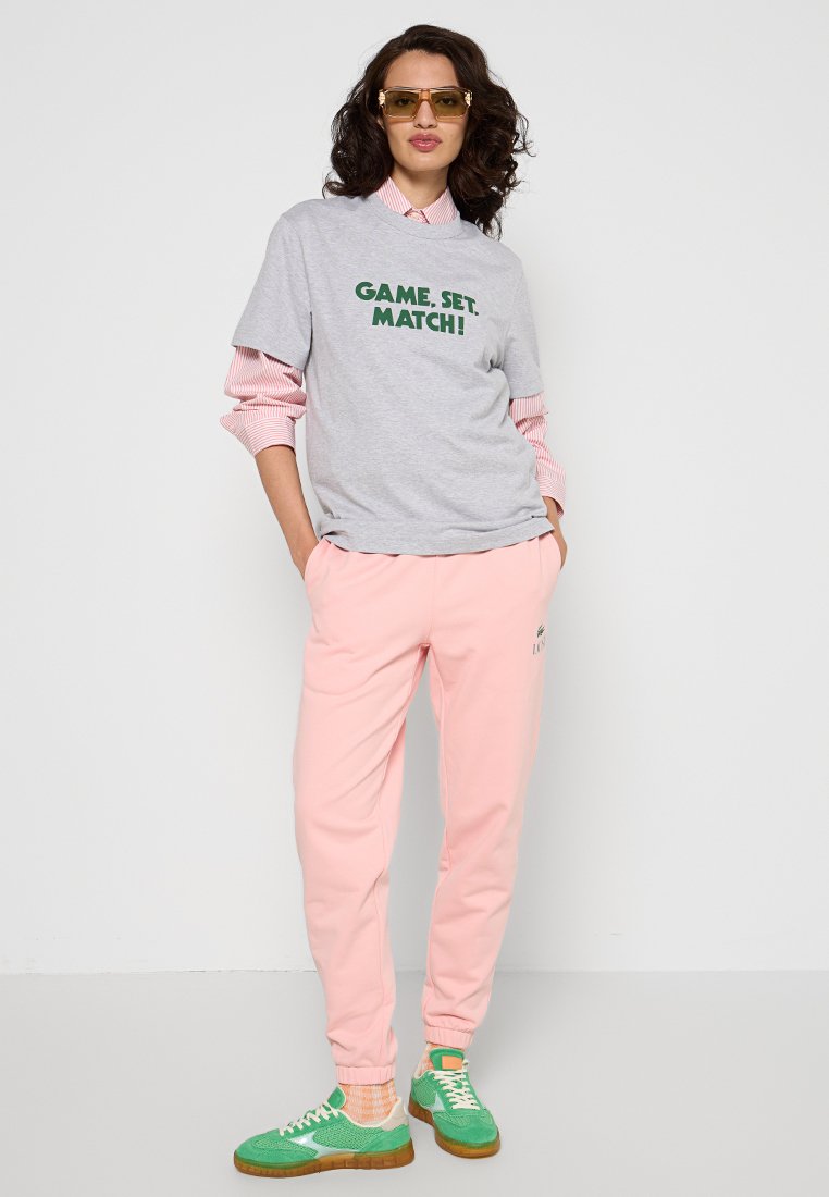 Graues T-Shirt mit "GAME, SET, MATCH!" in grüner Schrift, über einem pink-weißen gestreiften Hemd. Rosa Jogginghose und grüne Sneaker runden das Outfit ab.