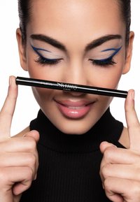 Penna per eyeliner nero tenuta orizzontalmente davanti alle labbra. Il modello sfoggia un trucco occhi azzurro intenso a effetto ala, toni neutri e una maglia nera a coste.
