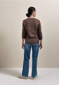 Brauner Strickpullover mit geripptem Saum, kombiniert mit hellblauen Schlagjeans. Das Modell steht mit dem Rücken zur Kamera vor einem neutralen Hintergrund.