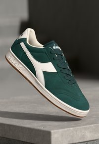 Green suede sneaker with white leather accents, textured gray rubber sole, and dark green laces. Features logo on tongue and side.