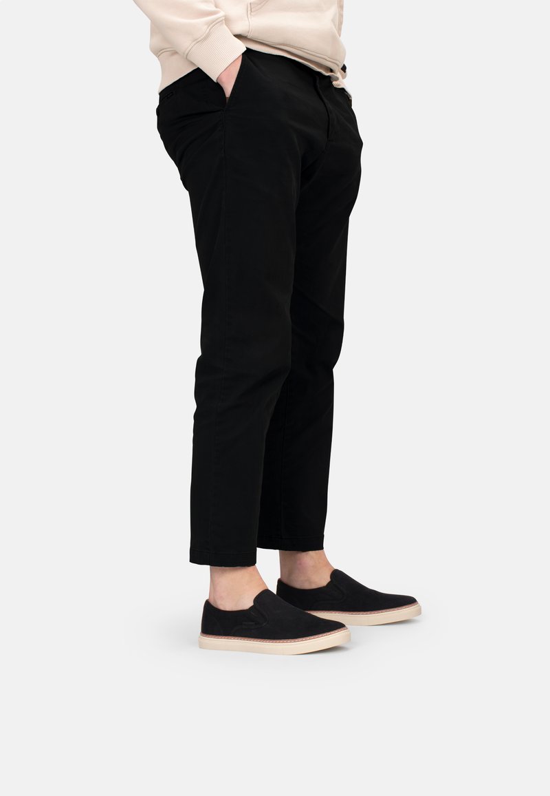 Schwarze Slipper aus Wildleder mit beigem Gummisohle, kombiniert mit schwarzen, schmal geschnittenen Hosen und einem hellen Kapuzenpullover.