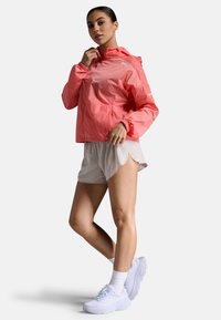 Lichte, gewatteerde jas met capuchon in felroze, gecombineerd met crèmekleurige shorts en witte sportschoenen. Glad textuur, casual ontwerp.