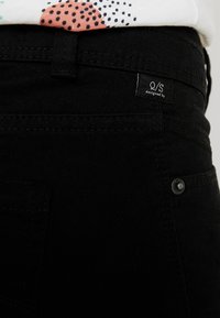 Jeans de denim negro con un primer plano de la cinturilla, que presenta una etiqueta cosida y un detalle de botón de metal.