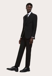 Schwarzer Anzug mit maßgeschneiderter Passform, bestehend aus einem einreihigen Sakko, passenden Hosen und polierten schwarzen Schuhen, getragen mit einem weißen Hemd und Krawatte.