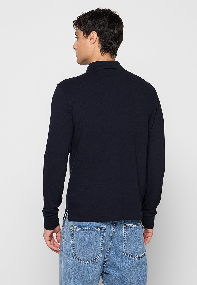 Hombre con una camisa polo negra de manga larga y vaqueros azul claro, de pie de espaldas sobre un fondo gris claro liso.