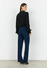Sort bluse med blonder ved ærmerne, parret med marineblå bukser. Outfittet har løs pasform og diskrete detaljer. Hæle fuldender looket.