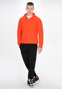 Lys orange fleece pullover med en kvart lynlås, parret med sorte bukser og sorte sneakers med røde og hvide detaljer.