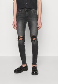 Jeans pretos justos com joelhos rasgados, feitos de denim. Apresentam um acabamento desbotado com bordas desfiadas. Usados com sapatos de couro pretos.