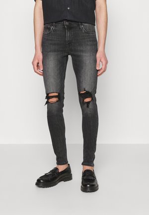 Jeans pretos justos com joelhos rasgados, feitos de denim. Apresentam um acabamento desbotado com bordas desfiadas. Usados com sapatos de couro pretos.