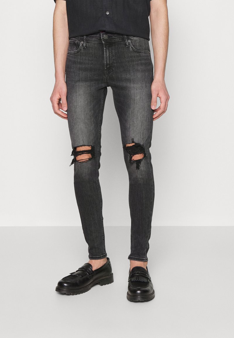 Jeans pretos justos com joelhos rasgados, feitos de denim. Apresentam um acabamento desbotado com bordas desfiadas. Usados com sapatos de couro pretos.