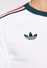 T-shirt de sport blanc avec un col côtelé vert, présentant des rayures rouges, blanches et vertes sur les épaules ainsi qu'un logo vert brodé.