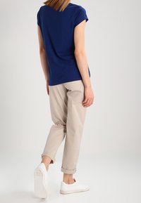 Person som bär en marinblå t-shirt, beige uppvikta byxor och vita sneakers, står med ryggen mot betraktaren på en neutral bakgrund.