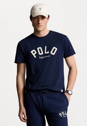 Camiseta de algodón azul marino con "POLO Ralph Lauren" en letras color crema, combinada con pantalones de chándal azul marino a juego y una gorra beige con un pequeño logotipo.