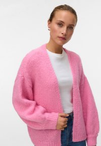 Ροζ πλεκτή ζακέτα με φαρδιά, μακριά μανίκια και ριμπ στην κάτω άκρη, φορεμένη πάνω από λευκό T-shirt. Το υφή του νήματος δημιουργεί μια μαλακή εμφάνιση.