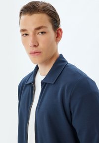 Chaqueta navy con cremallera y textura acanalada, llevada sobre una camisa blanca. La cremallera cuenta con un acento metálico.