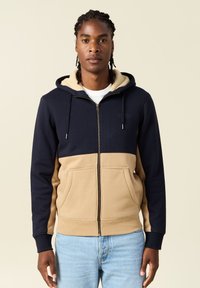 Felpa con cappuccio e zip, con parte superiore blu navy e parte inferiore color beige, dotata di cappuccio foderato in morbido tessuto, due tasche frontali e manicotti a costine.