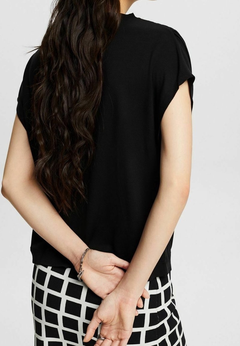 Femme aux longs cheveux ondulés foncés portant un haut noir à manches courtes et un pantalon à carreaux noir et blanc, mains jointes dans le dos.